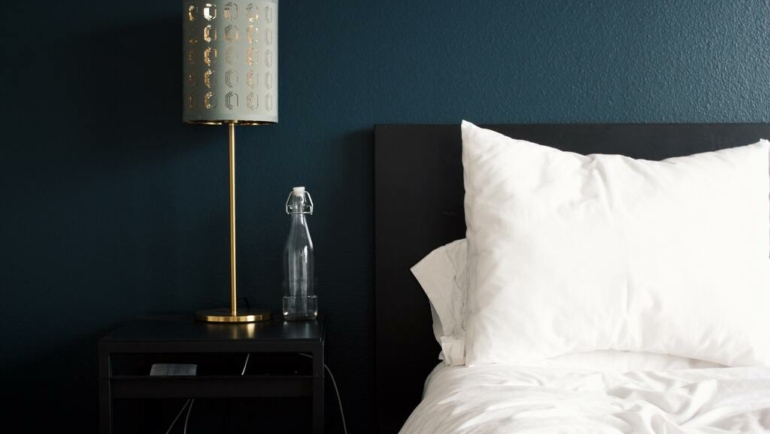 cama de hotel com lençõis e travesseiro brancos, mesa de cabeceira ao lado com uma garrafa de água e luminária