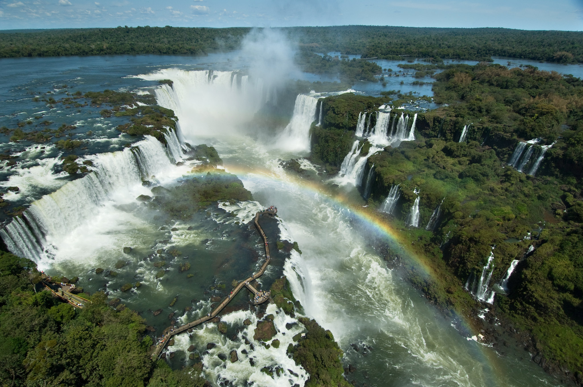Vista das Cataratas do Iguaçu com passarela de turistas e arco-íris ao fundo